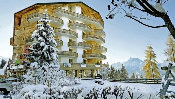 Un immeuble à plusieurs étages doté de balcons, entouré d’arbres couverts de neige et avec des montagnes en arrière-plan.