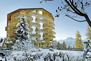 Un edificio de varios pisos con balcones, rodeado de árboles nevados y montañas al fondo.
