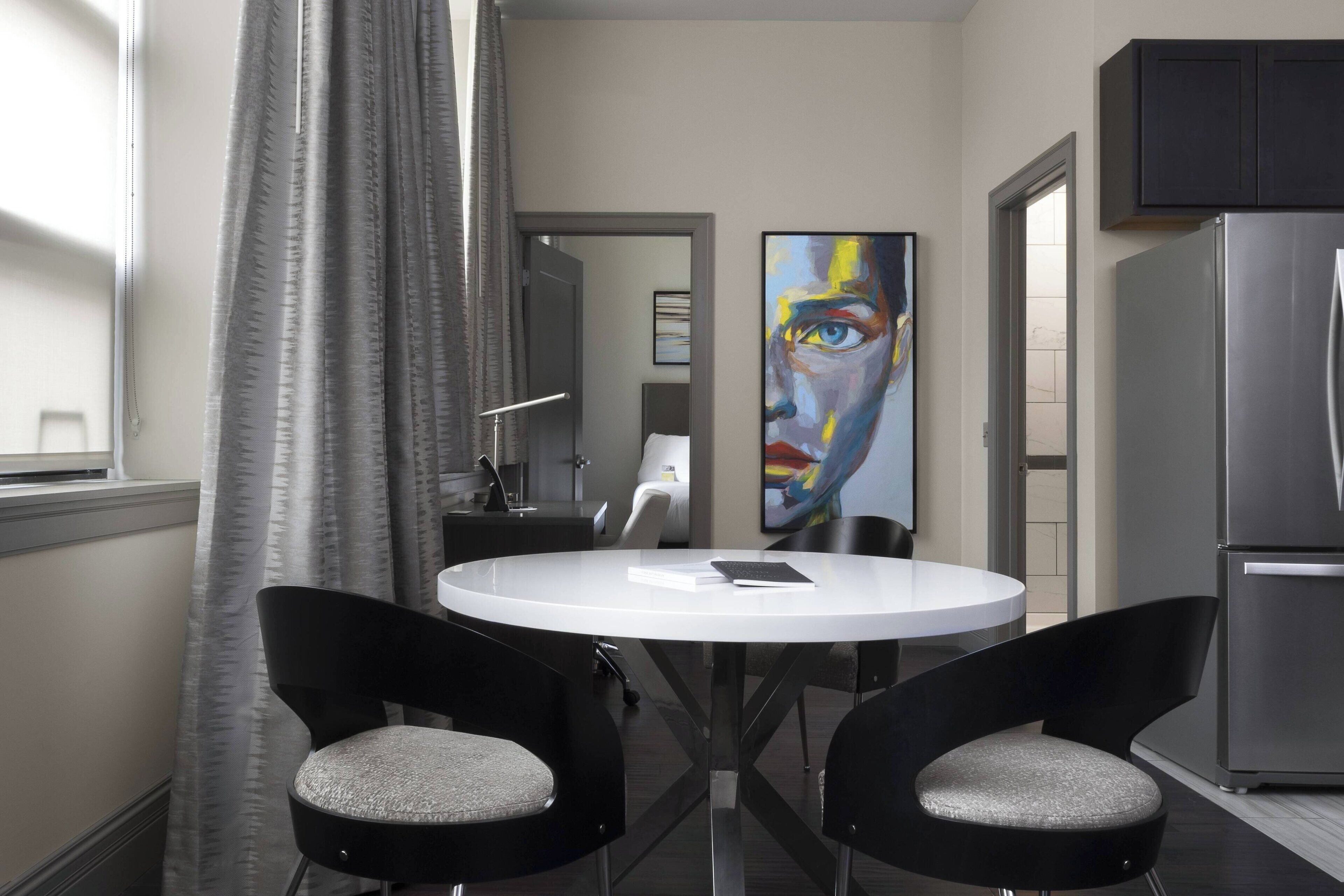 A modern kitchen with a stainless steel refrigerator, a white dining table with two black chairs, and a large abstract painting on the wall.