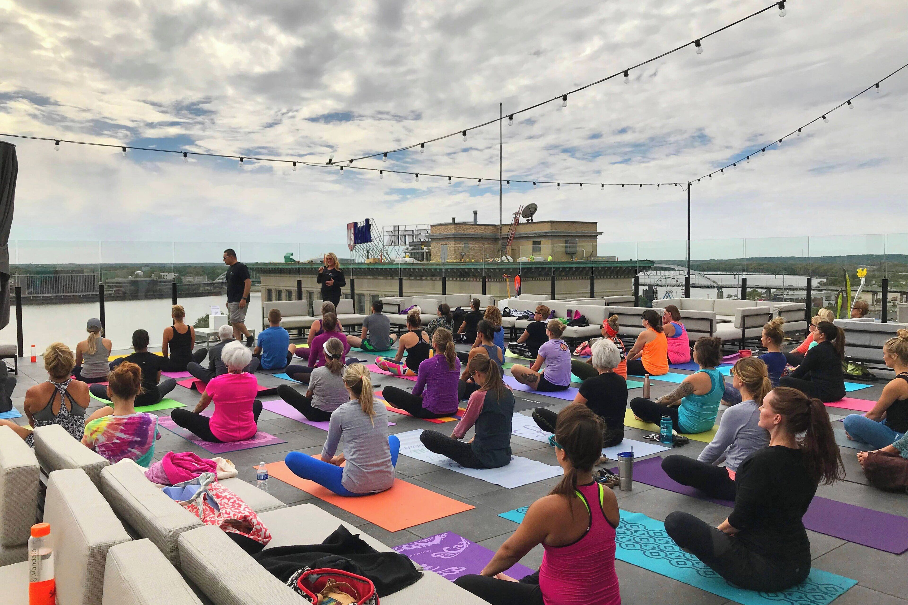 A group of people sitting on yoga mats outdoors on a rooftop, with a cityscape in the background.