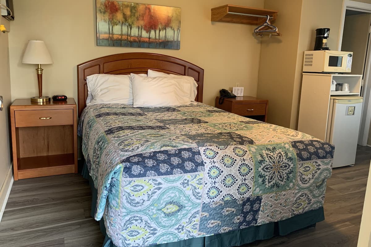 A neatly made bed with a patterned quilt, a bedside lamp, a nightstand, a wooden shelf, a coffee maker, and a refrigerator.