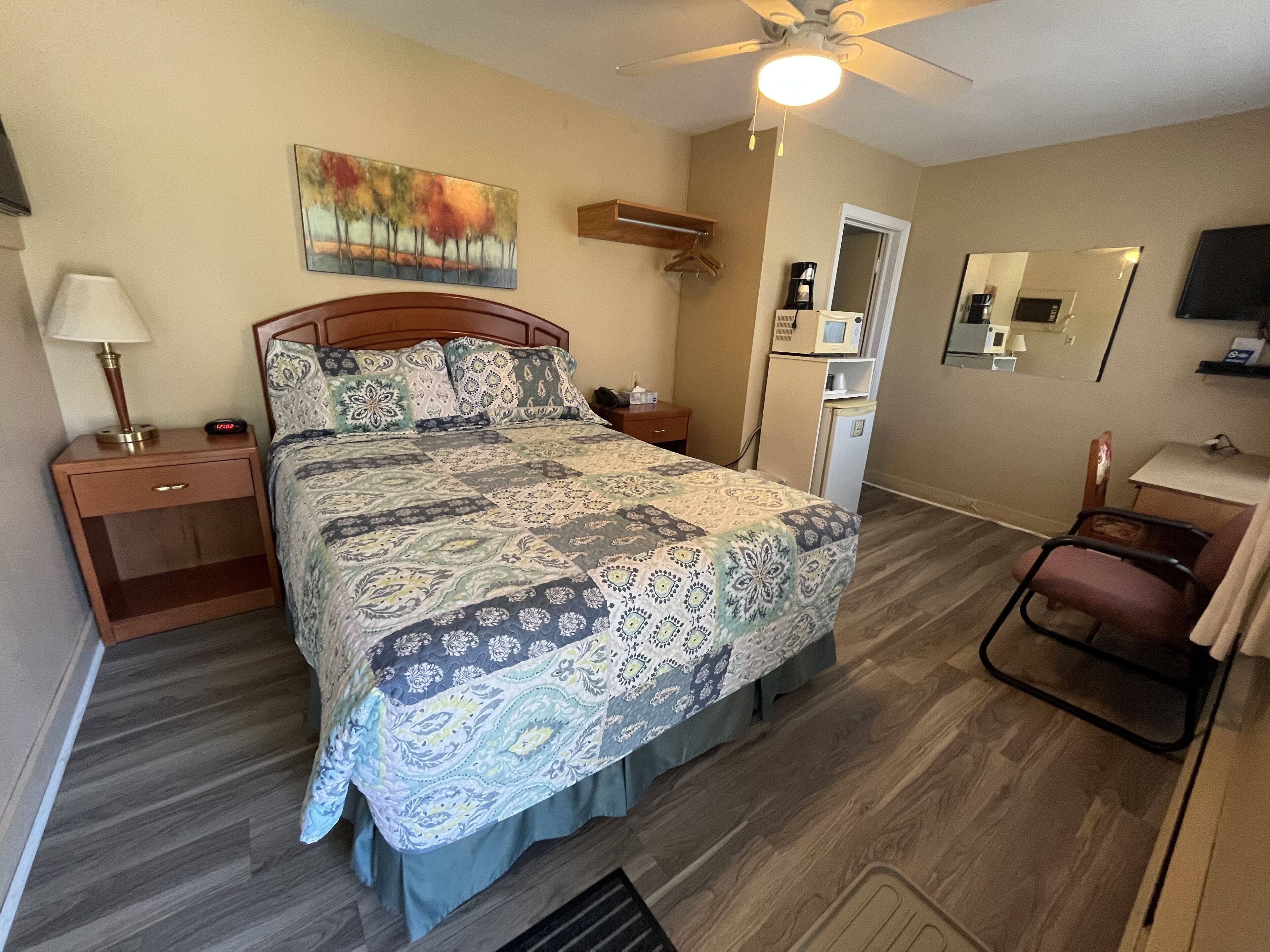 A bedroom with a bed, bedside tables, a desk, a chair, a ceiling fan, a microwave, a refrigerator, a painting, and a mirror.