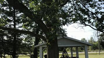 Un kiosque avec une table de pique-nique et une balançoire, situé dans un espace verdoyant et arboré.