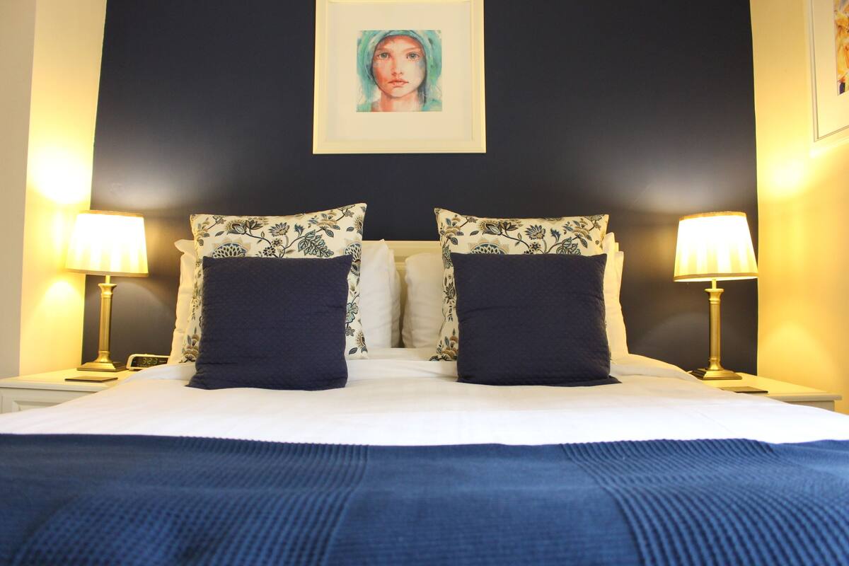Un lit bien fait, recouvert d’une couverture bleue, avec deux oreillers assortis et une œuvre d’art encadrée au-dessus.