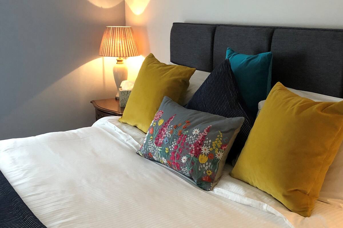 Un lit bien fait, avec du linge de lit blanc, une variété d’oreillers colorés et une lampe sur une table de chevet.