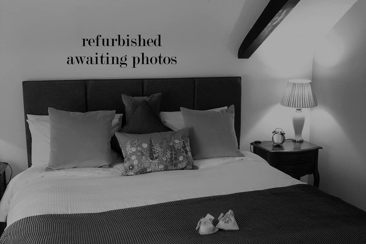 Une chambre rénovée comprenant un lit, des oreillers, une table de chevet avec une lampe, et une paire de chaussures posée sur le lit.