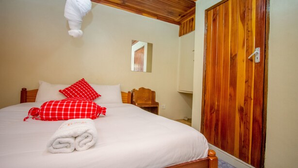 Een net opgemaakt bed met witte lakens en rode geruite kussens, een houten garderobe en een klein nachtkastje met spiegel.