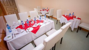 Een hotelrestaurant met tafels voor vier personen, elk voorzien van een witte tafelkleed en een rode placemat, met waterflessen, glazen en bestek.