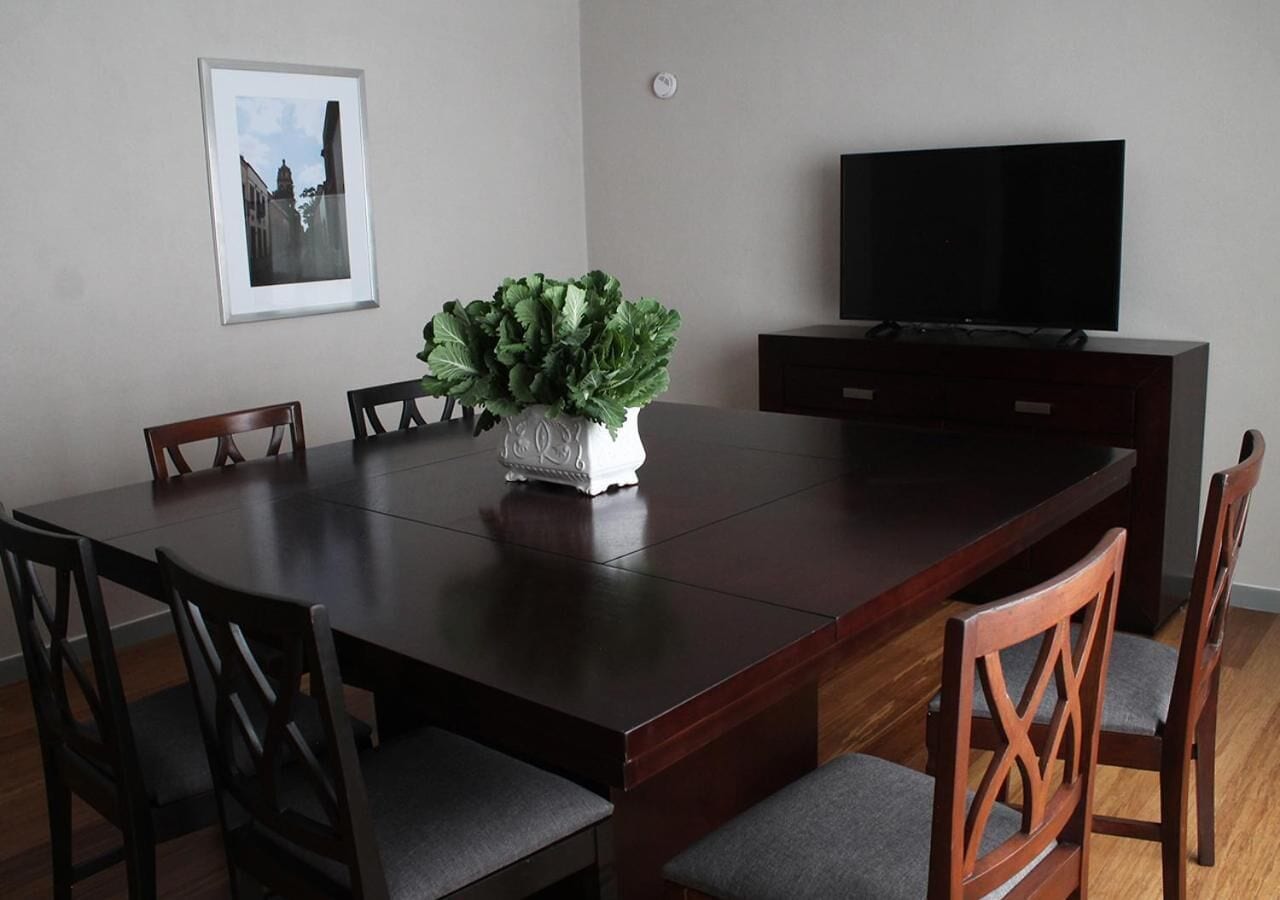 Un comedor con una mesa de madera oscura, seis sillas, un jarrón blanco con follaje verde, un cuadro en la pared y un televisor sobre un mueble de madera oscura.