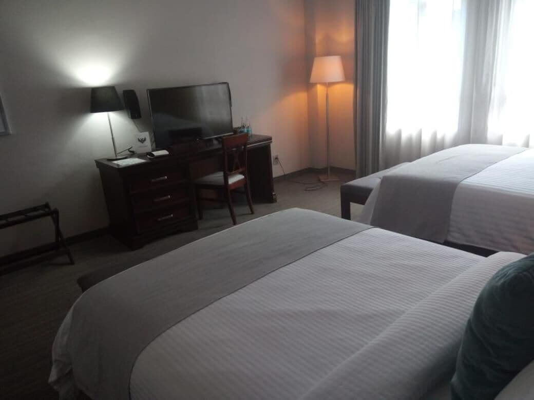 Habitación de hotel con dos camas, un escritorio con televisor, una lámpara y una ventana con cortinas.