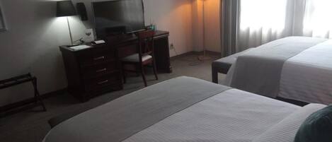 Ein Hotelzimmer mit zwei Betten, einem Schreibtisch, einem Fernseher, einer Lampe und einem Fenster mit Vorhängen.