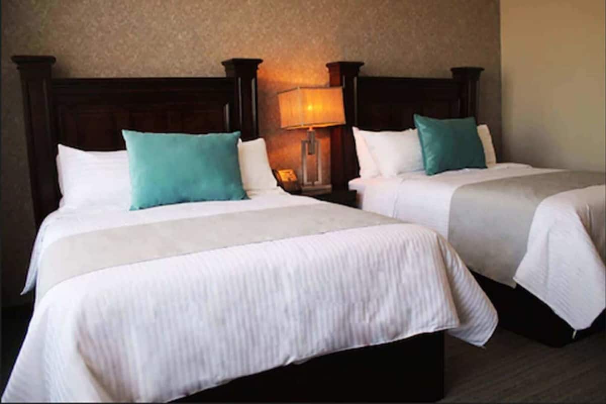Two double beds with white linens and teal pillows, a nightstand with a lamp, and a textured wall.