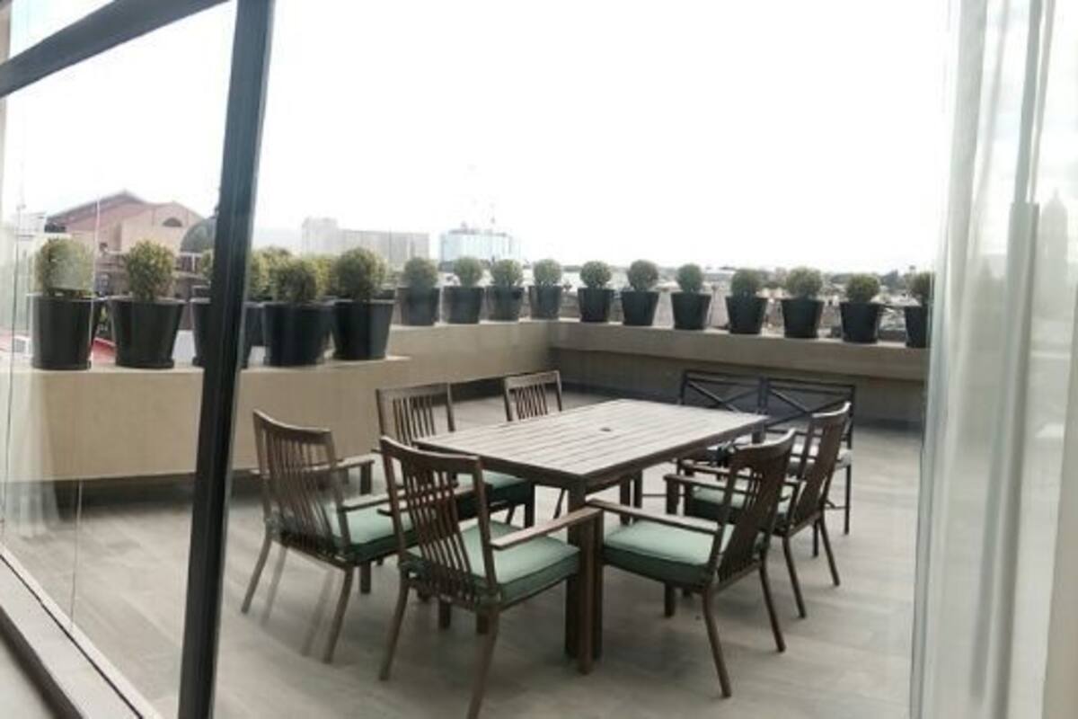 A hotel room with a bed, two armchairs, a table with chairs, a balcony with potted plants, and a view of the city.