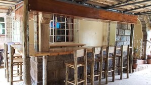 A rustic wooden bar with a stone wall backdrop, wooden stools, and a window with metal bars. - Sahara Gardens Kericho (Kericho)