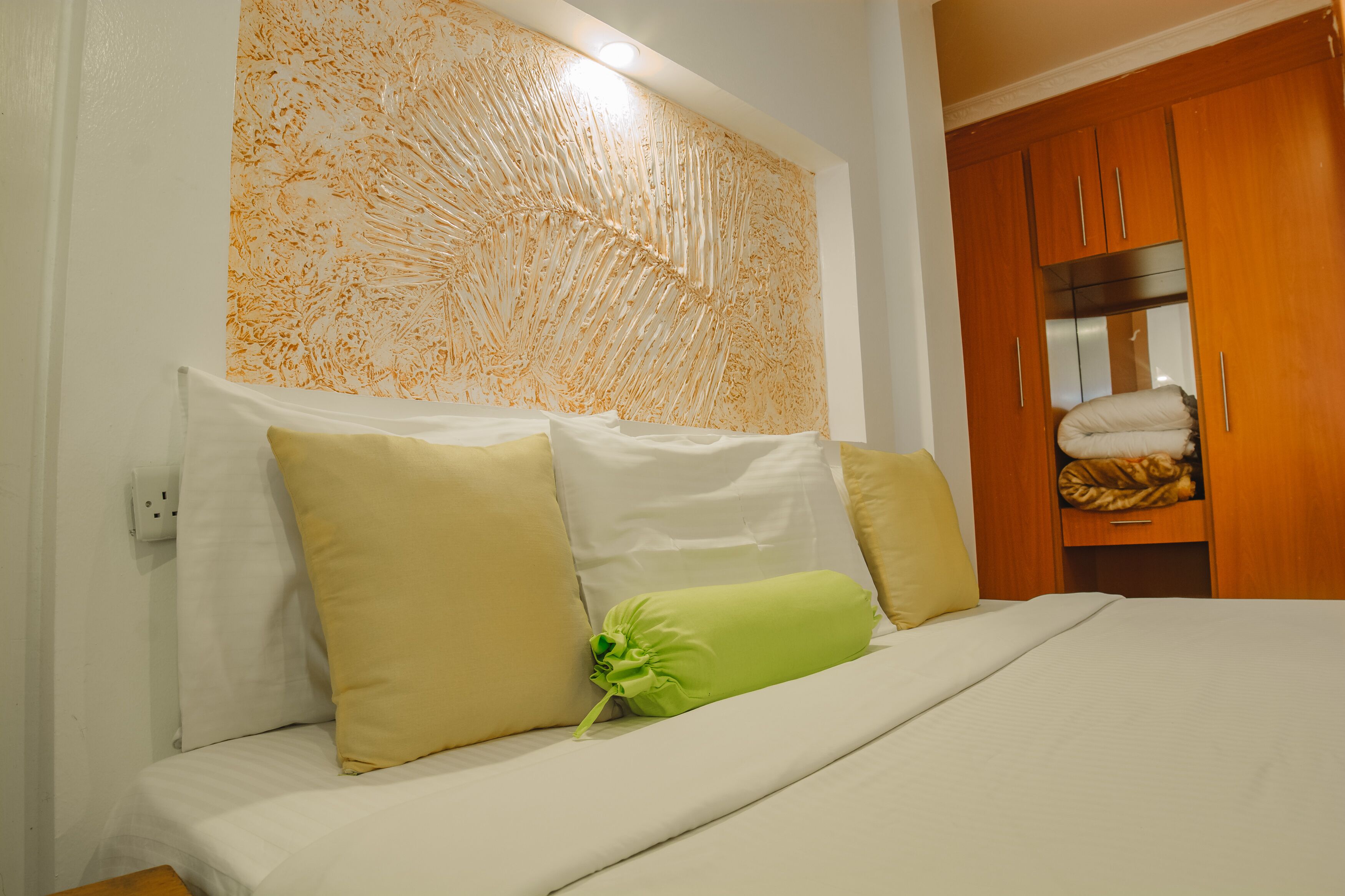 Una cama bien tendida con ropa de cama blanca y beige, una almohada verde y una pared con diseño texturizado.