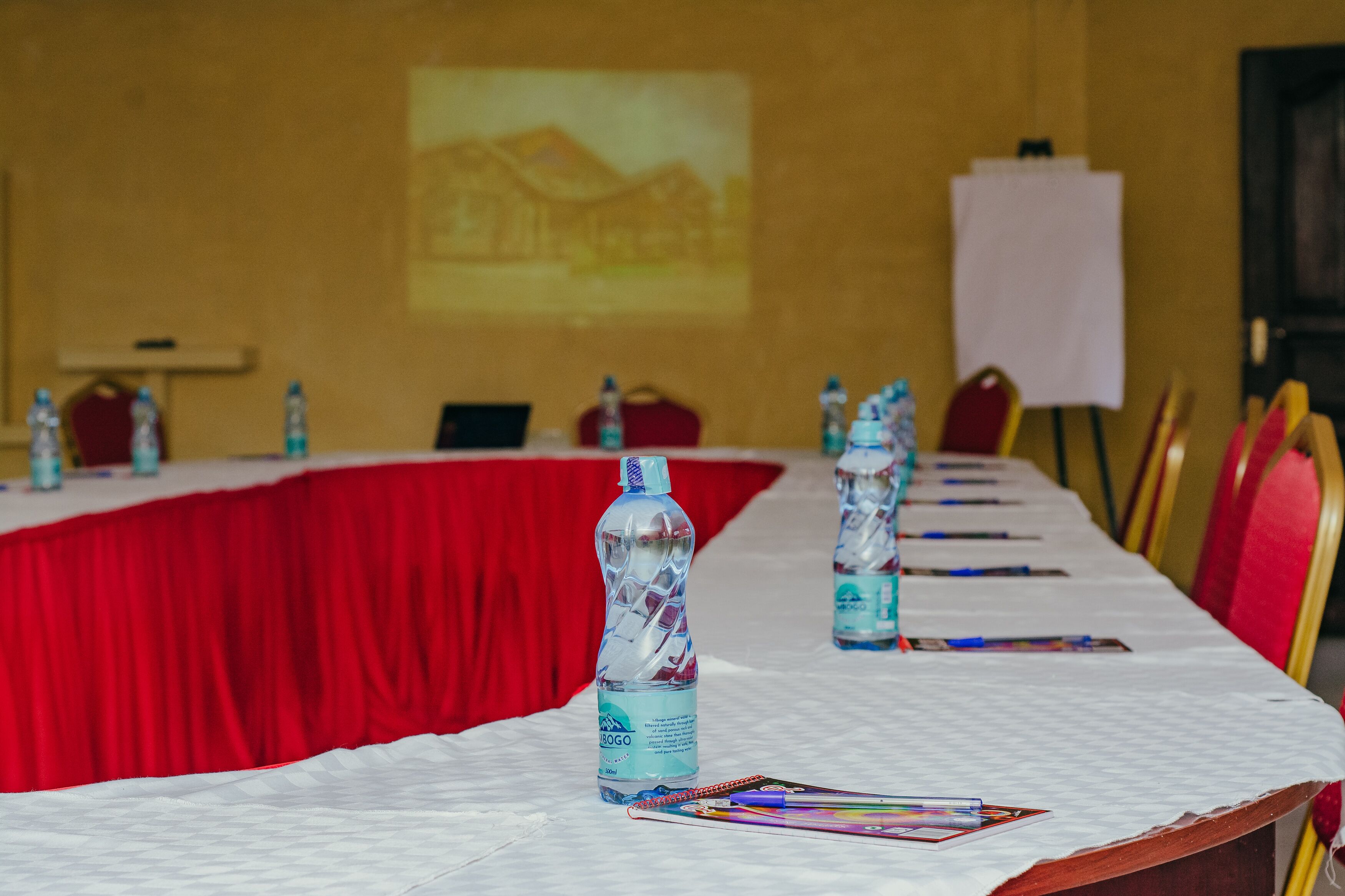 A conference room with a long table, red curtains, water bottles, and a projector screen on the wall.