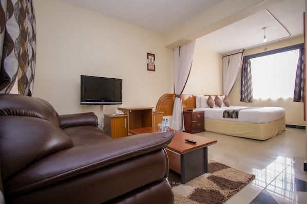 A hotel room with a leather sofa, a desk with a telephone, a bed with pillows, a TV, and a window with curtains. - Emboita Hotel (Nakuru)
