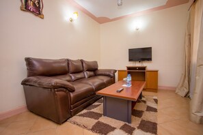 A living room with a brown leather sofa, a wooden coffee table, a television on a stand, and a patterned rug. - Emboita Hotel (Nakuru)