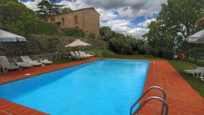 Une piscine rectangulaire avec un escalier en métal, entourée d’une terrasse carrelée de tuiles rouges, de chaises longues blanches et d’un mur de pierre, avec un bâtiment visible en arrière-plan.