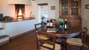 Un coin repas avec une cheminée, des chaises en bois, une table ronde ornée de bouteilles de vin et d’un livre, ainsi qu’un buffet en bois doté de portes en verre.
