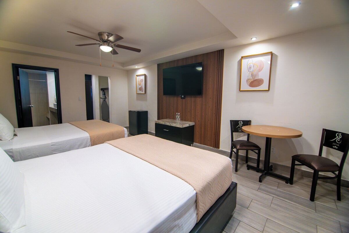 Une chambre d’hôtel avec deux lits, un ventilateur de plafond, une télévision, une petite table avec des chaises et un mur recouvert de panneaux en bois.