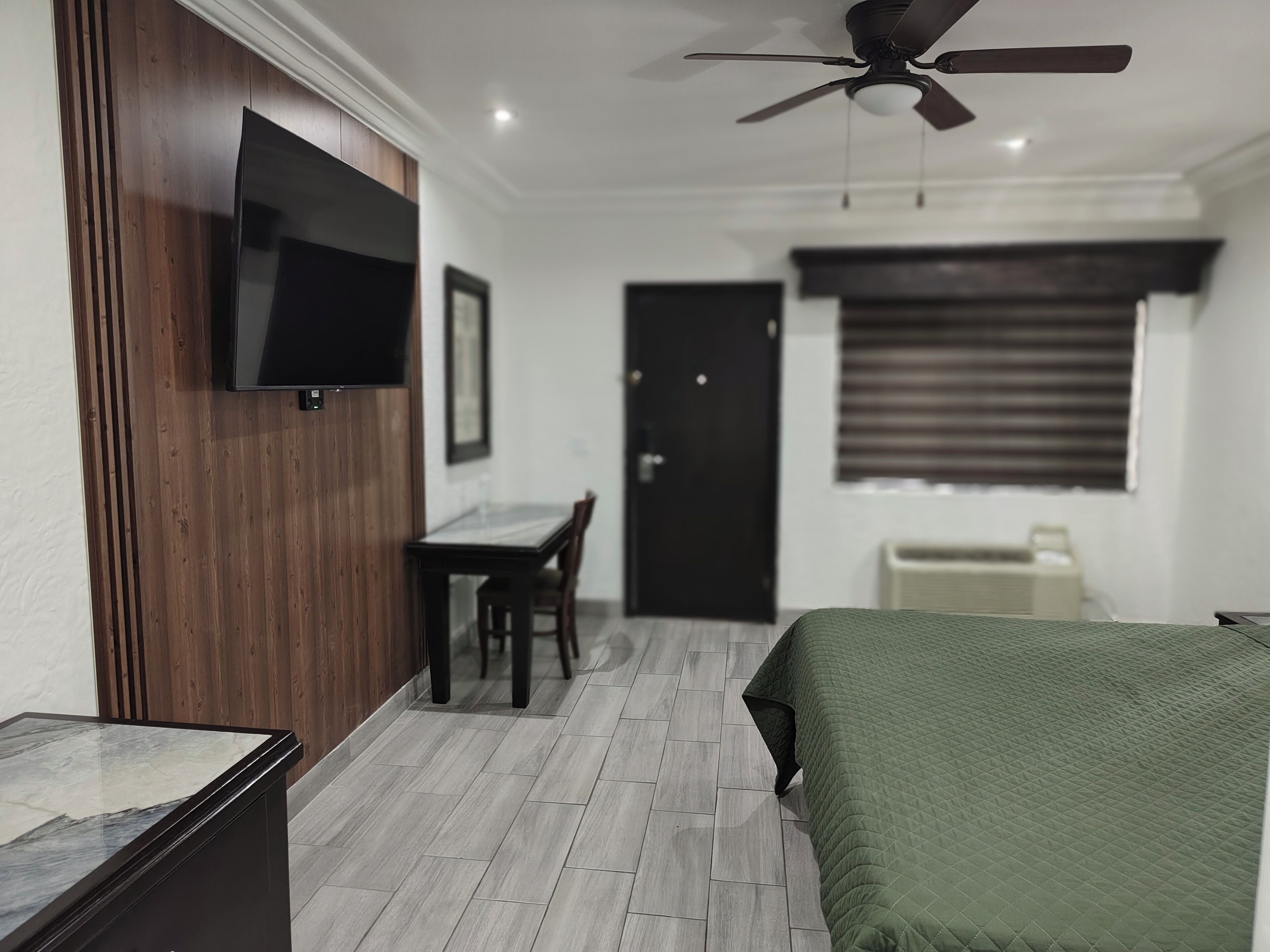 Habitación de hotel con cama, escritorio, silla, televisor montado en un panel de madera y ventilador de techo.
