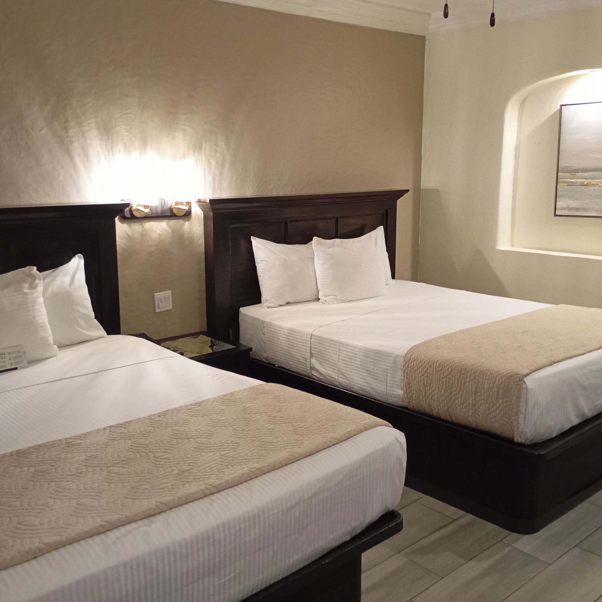 Een hotelkamer met twee bedden, elk voorzien van witte lakens en een donker hoofdeinde. Daarboven hangt een wandlamp en aan de muur een lijst met een afbeelding.