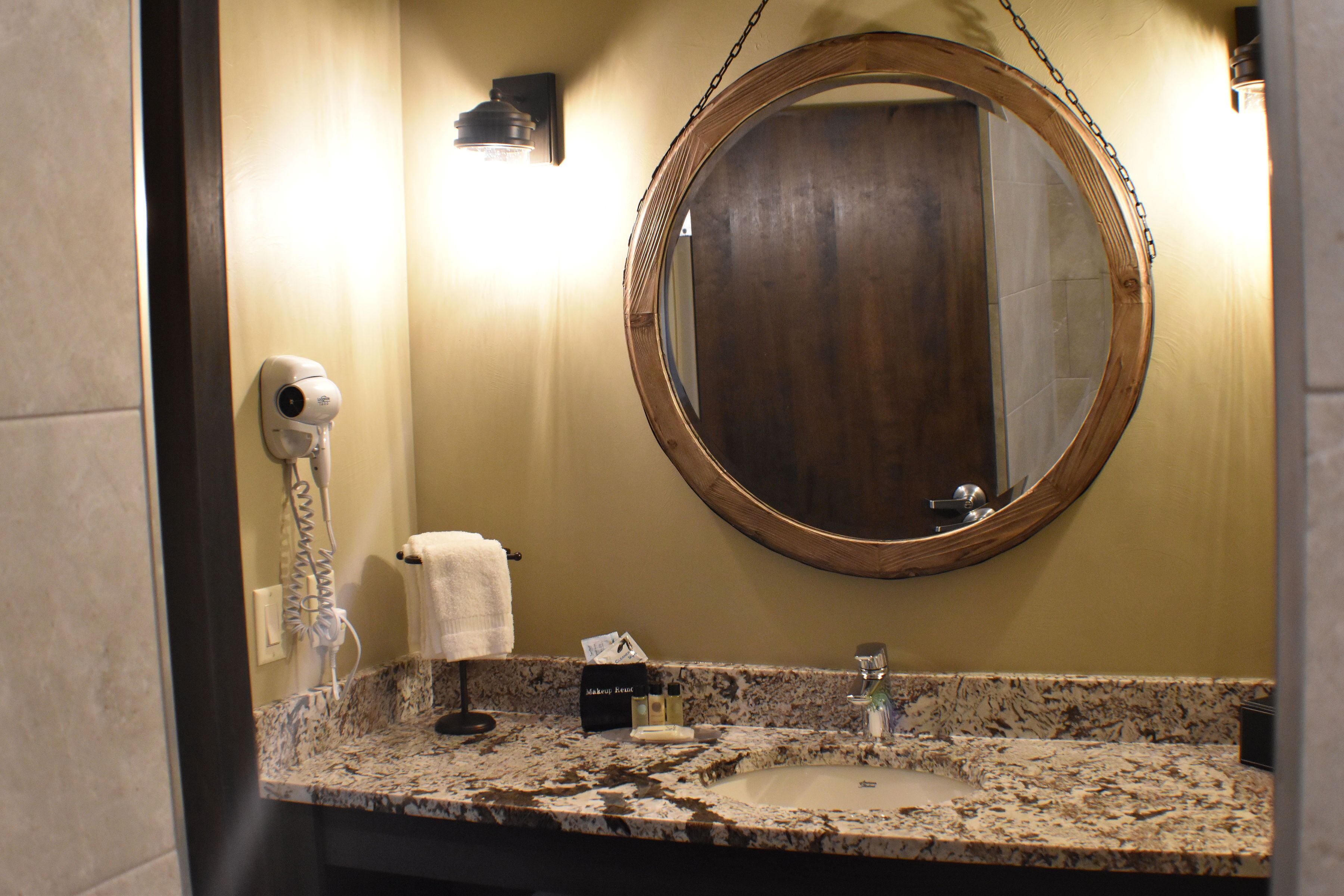 Une salle de bain dotée d’un plan de toilette en granit, d’un miroir rond, d’un porte-serviettes et d’un sèche-cheveux.
