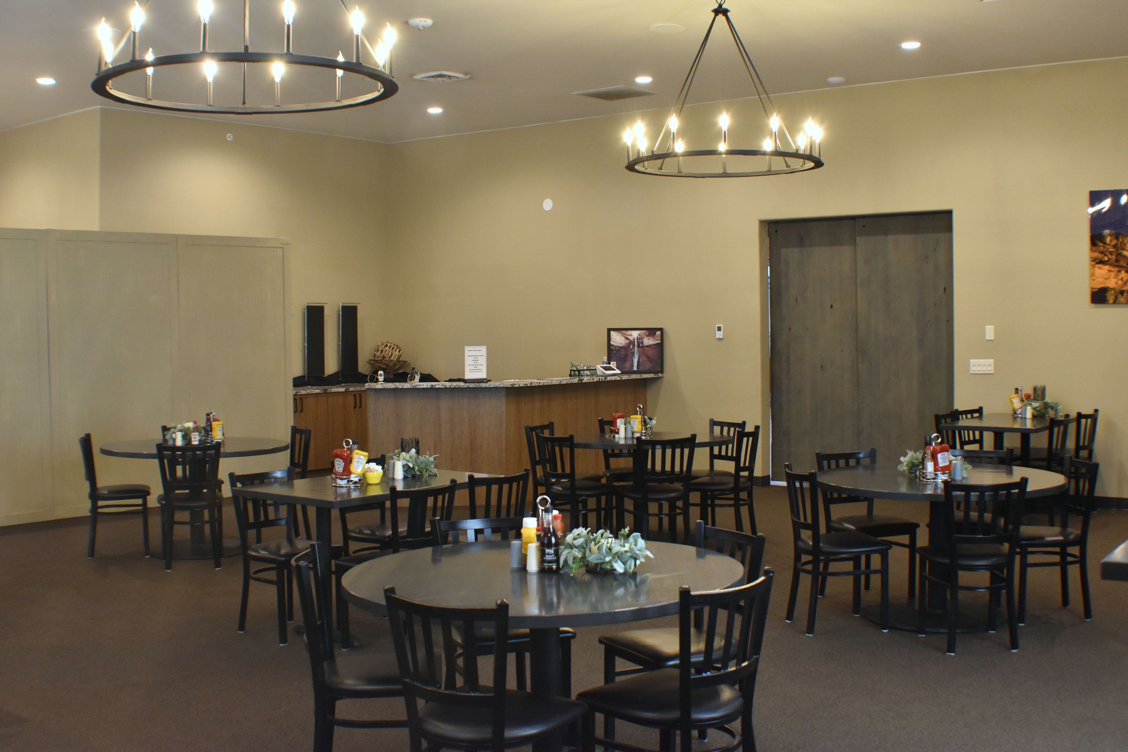A hotel dining area with multiple round tables and chairs, a bar counter, and two chandeliers.