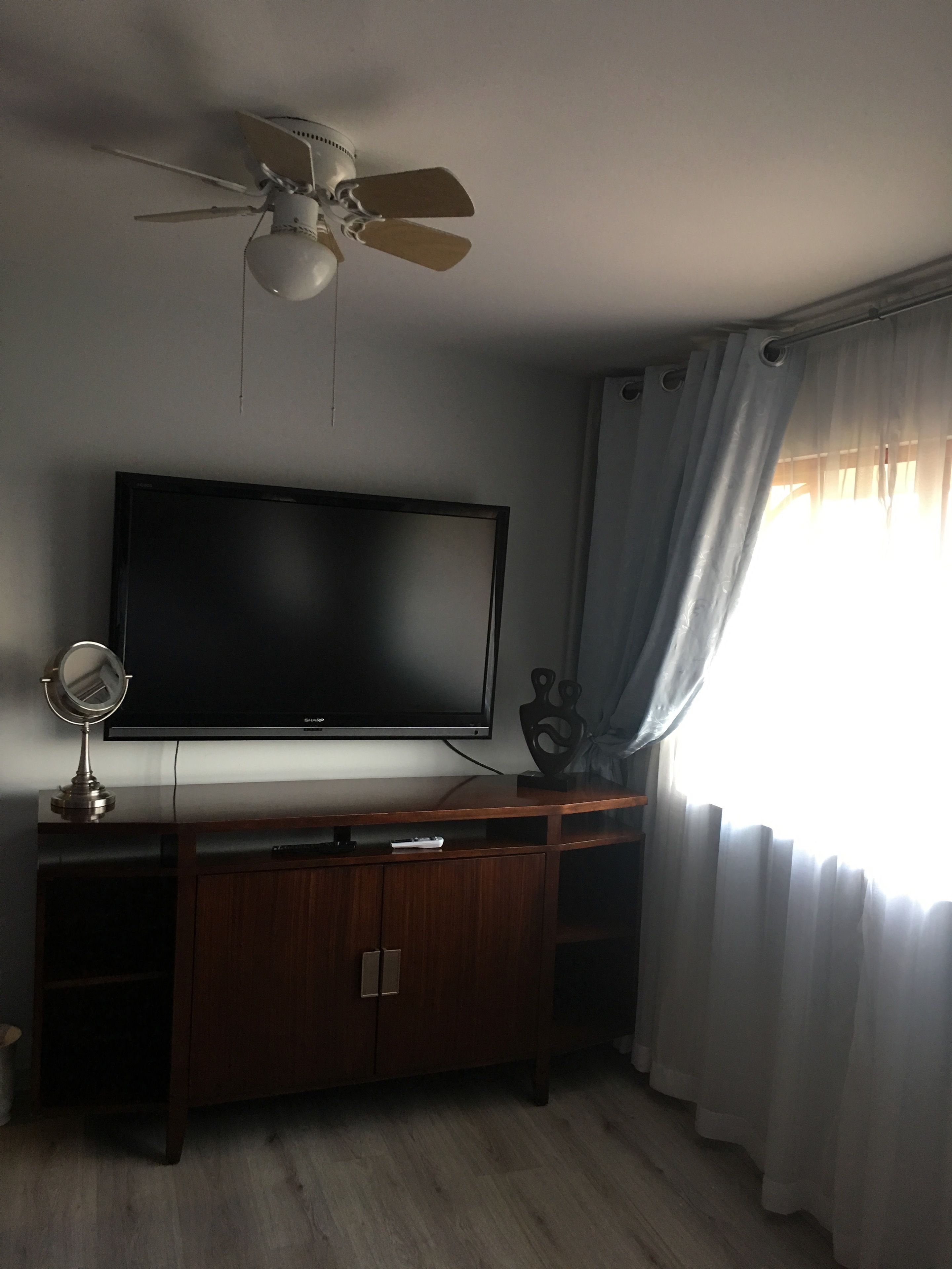 Une pièce dotée d’un ventilateur de plafond, d’un téléviseur à écran plat posé sur un meuble en bois, d’un petit miroir rond et d’une fenêtre ornée de rideaux légers.
