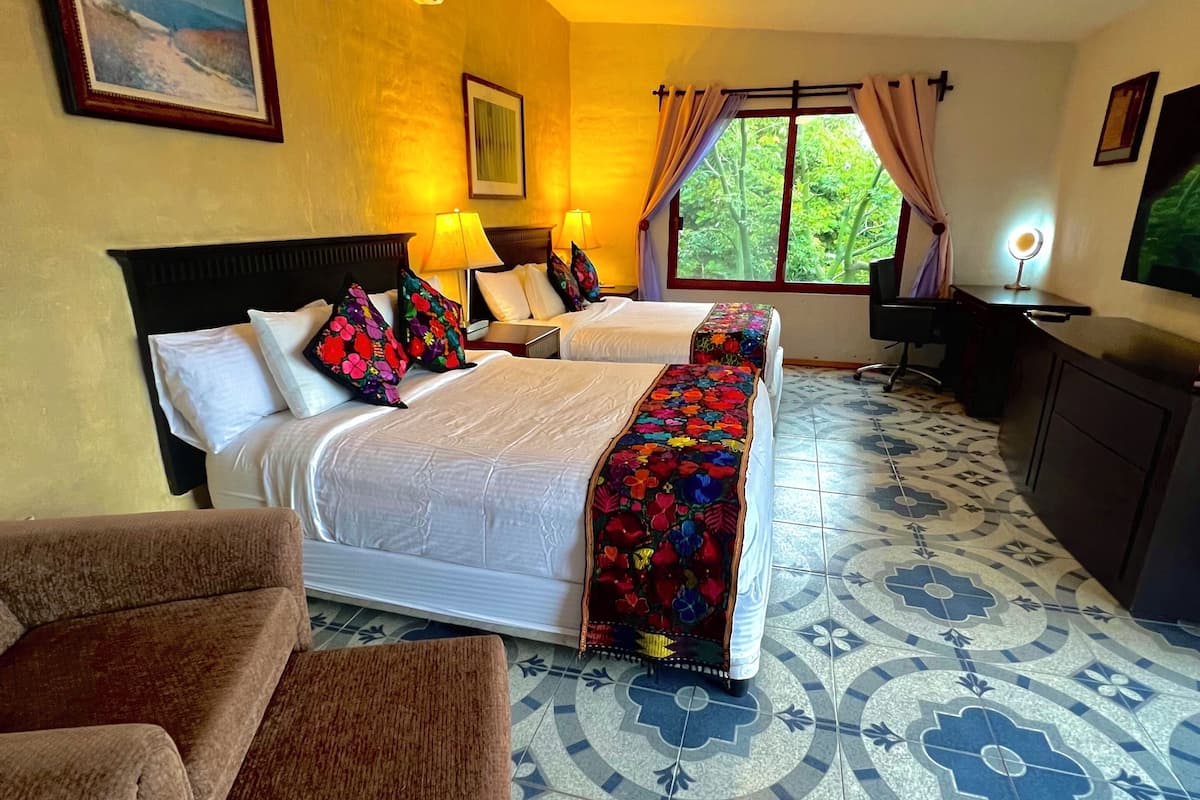 A hotel room with two beds, a ceiling fan, a desk with a chair, a TV, and a window with curtains.