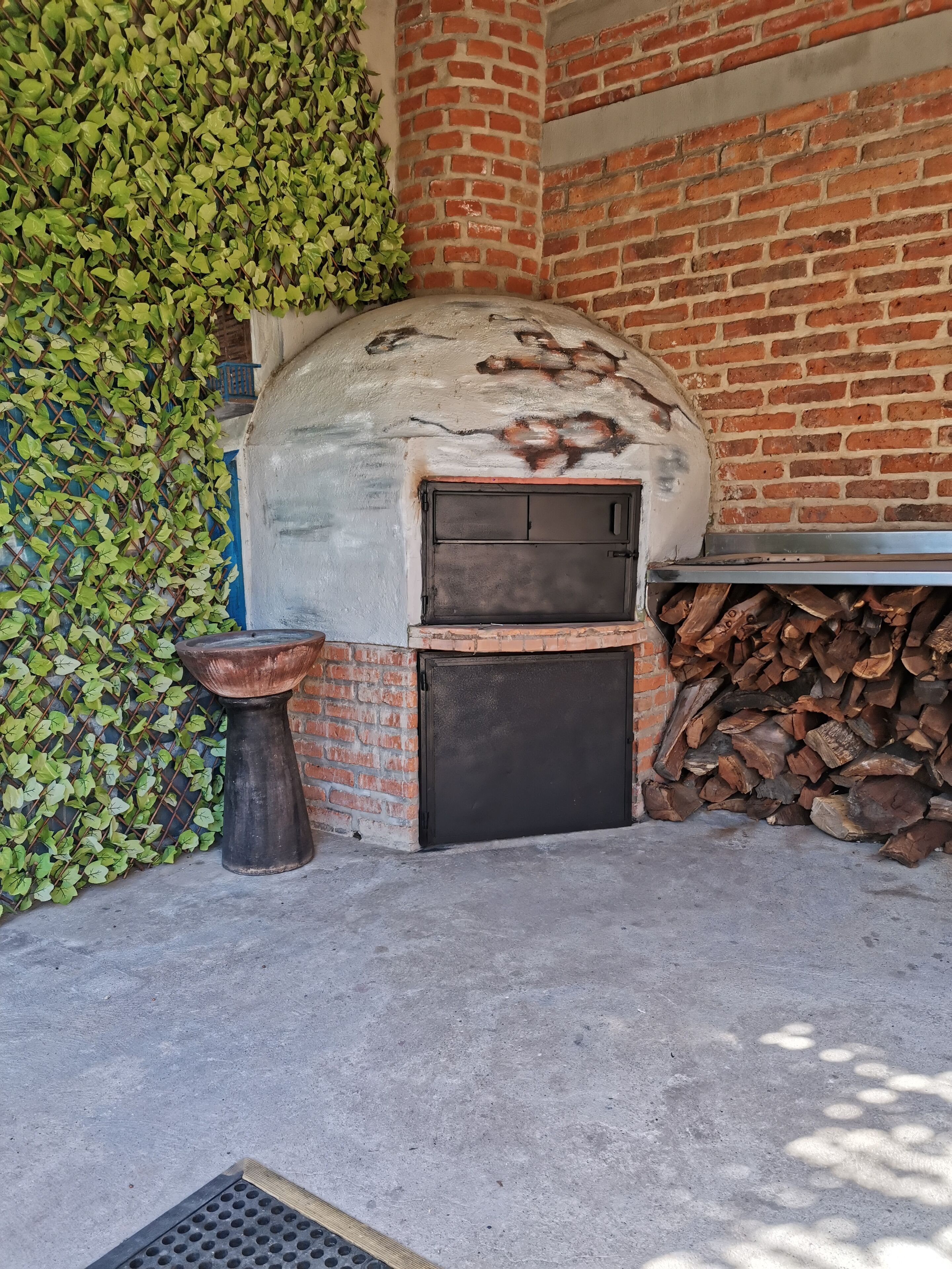Horno de ladrillo con puerta metálica, una bacha de piedra y una pila de leña junto a una pared de ladrillo con follaje verde.