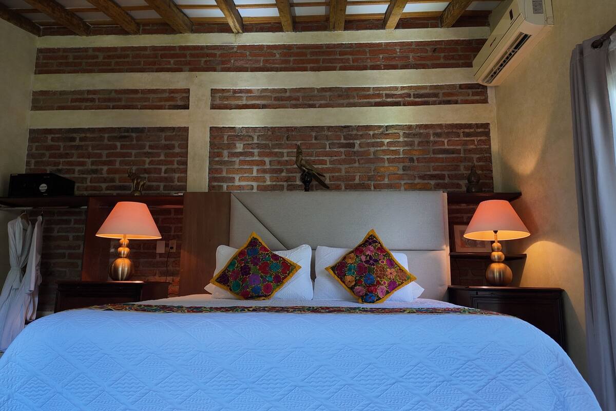 A hotel room with a brick wall, a bed with white bedding and colorful pillows, two bedside lamps, and a window with curtains.