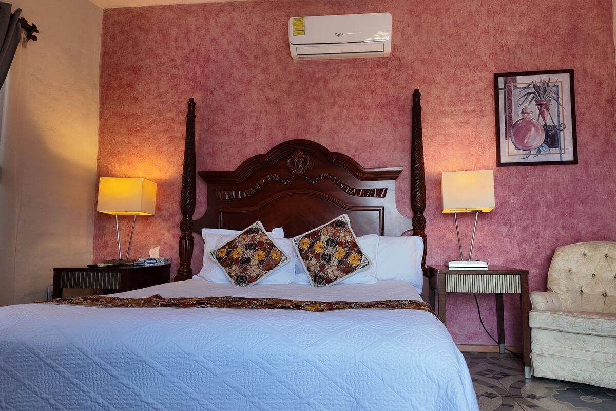A hotel room with a large bed, two bedside tables with lamps, a chair, an air conditioner, and a framed picture on the wall.