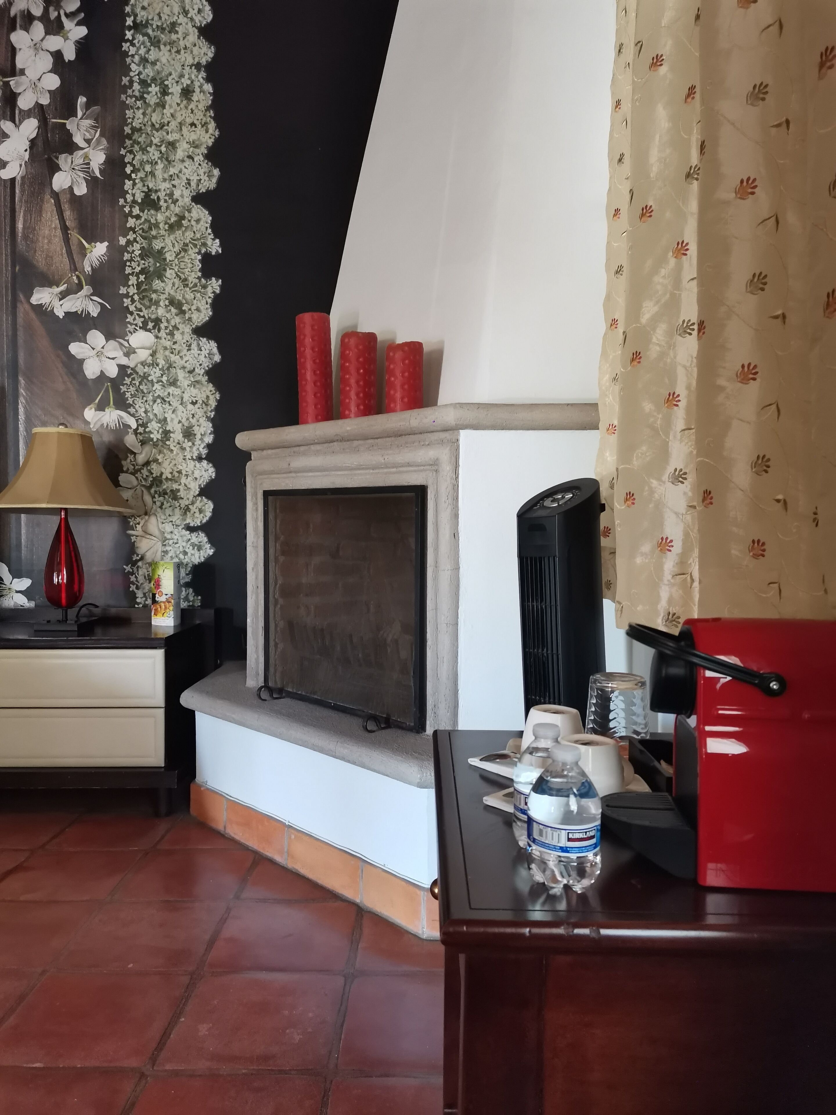 Una habitación con chimenea, una cafetera roja, una botella de agua y una lámpara sobre una mesita.