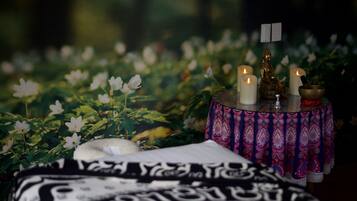 Una habitació amb un llençol estampat, una taula petita amb espelmes i una estàtua de Buda, i un fons de flors blanques.