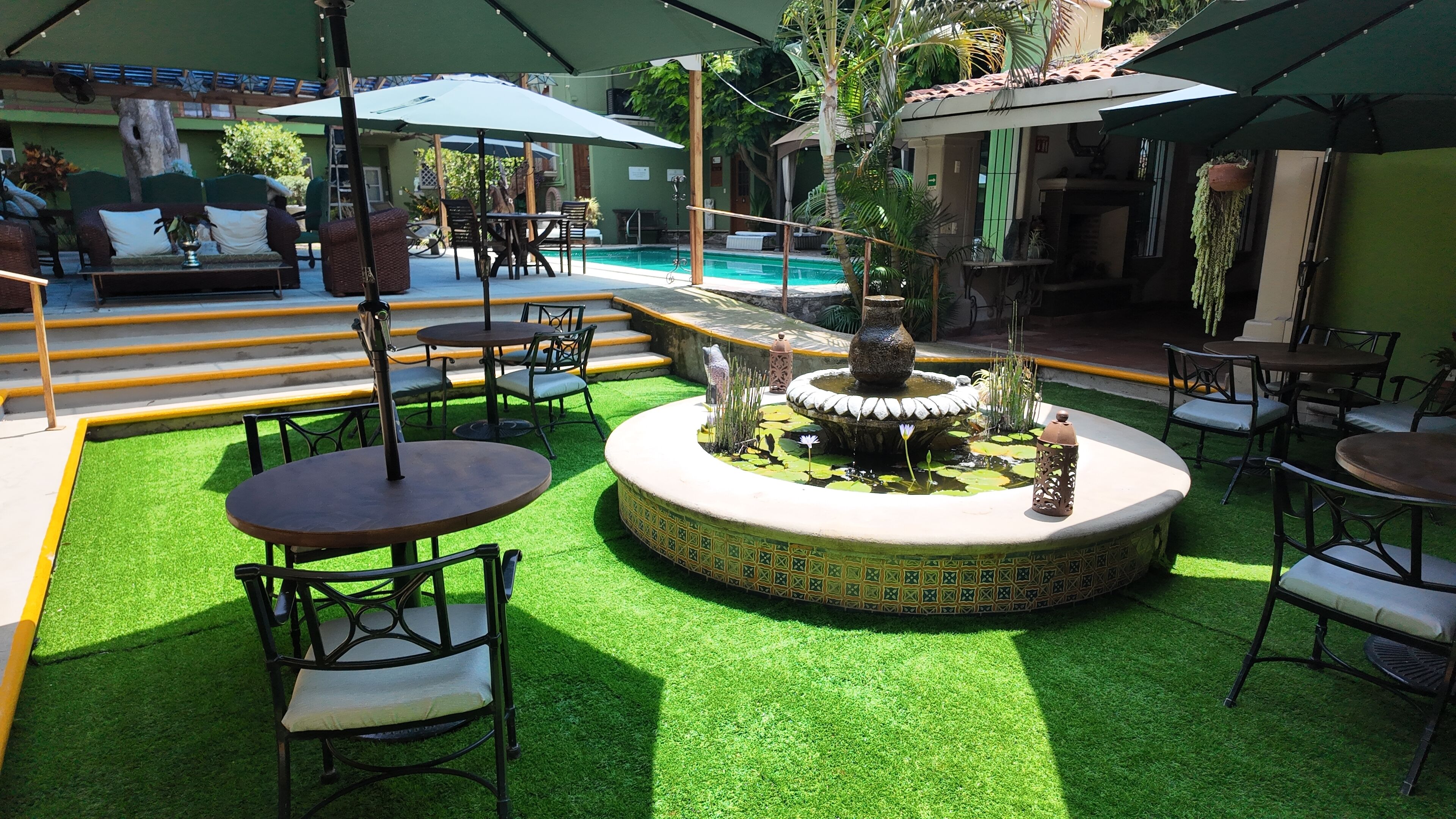 Zona junto a la piscina con césped artificial, una fuente central y mobiliario de exterior.