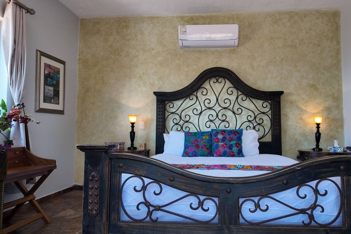 A bedroom with a bed, two bedside lamps, an air conditioning unit, a framed picture, and a wooden nightstand.
