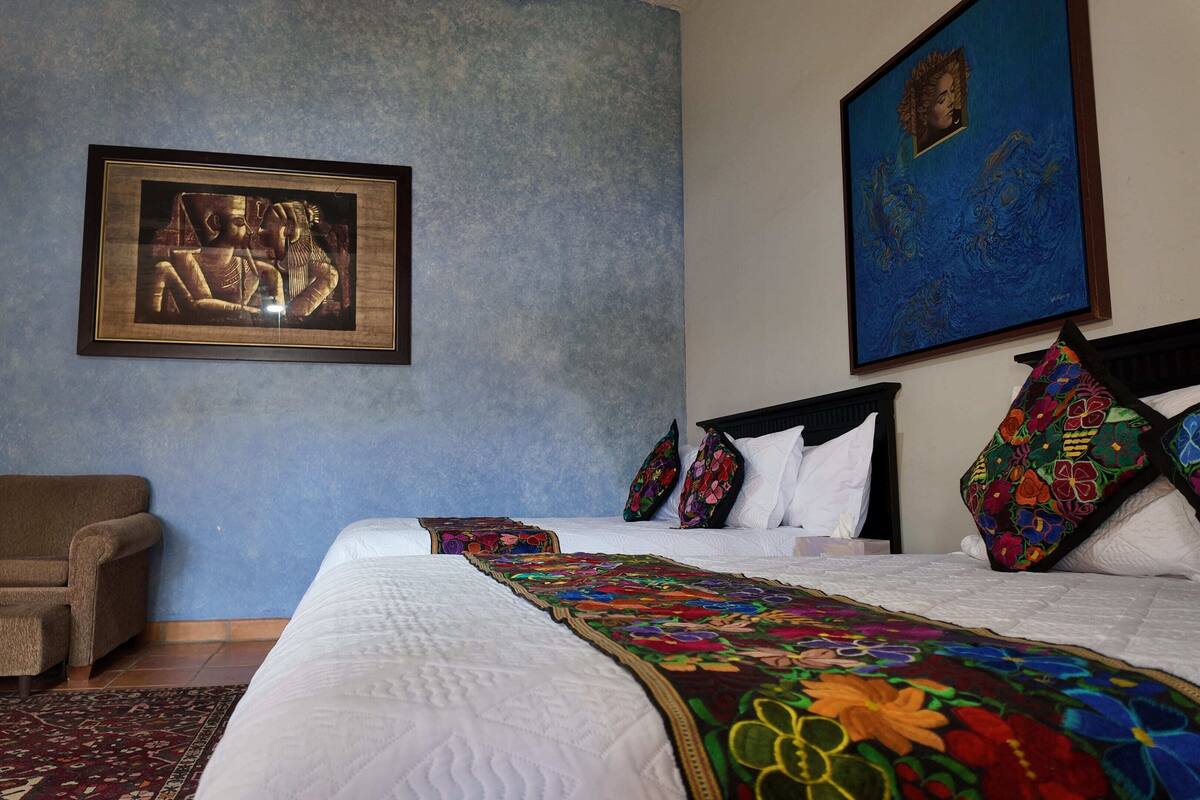 A hotel room with a bed, a chair, a colorful quilt, and two framed artworks on the wall.