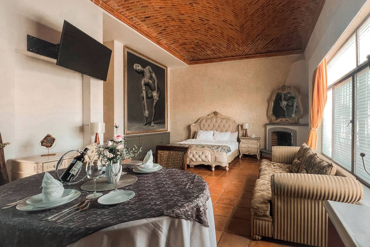 A hotel room with a bed, a sofa, a dining table set for two, and a fireplace.