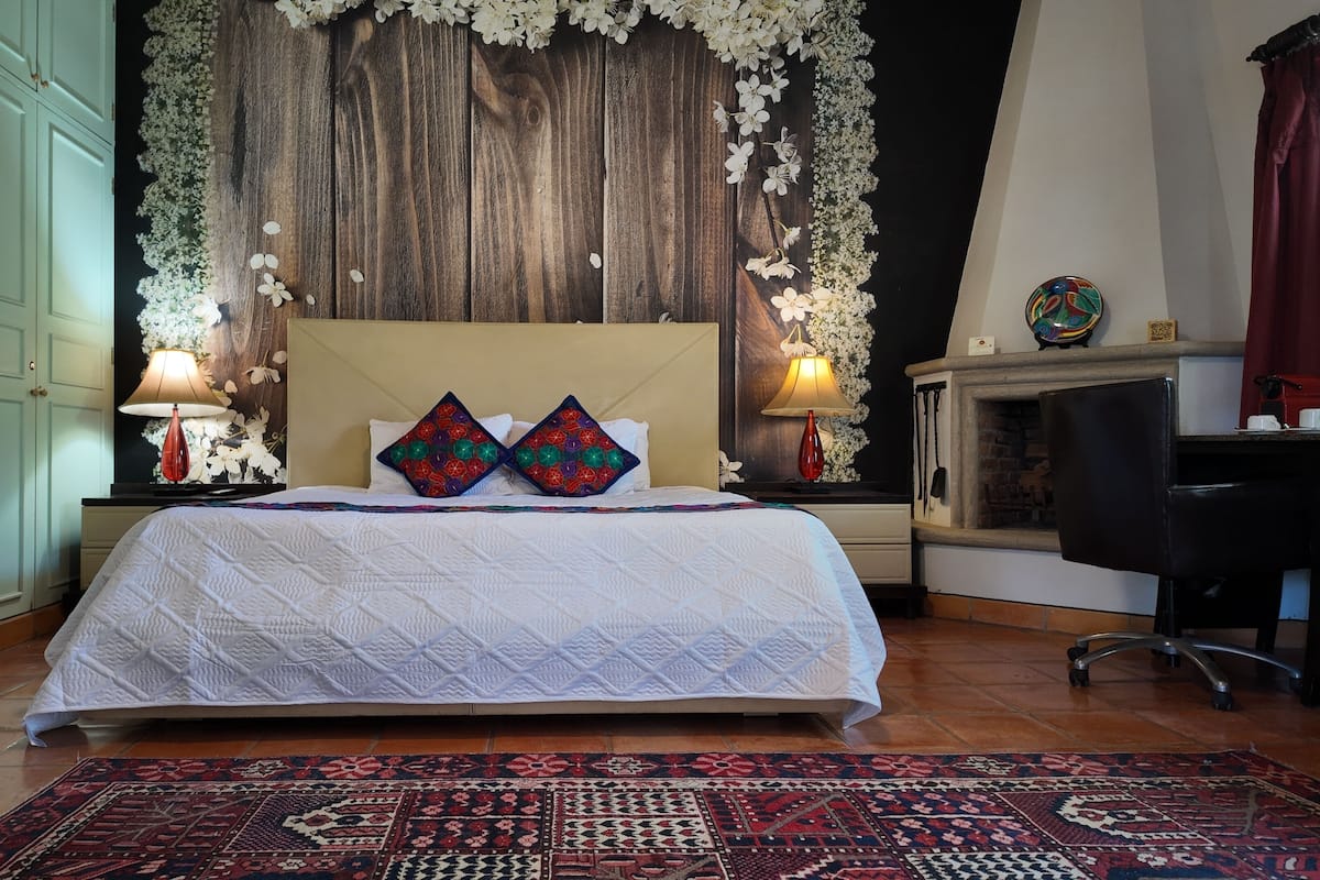A bedroom with a large bed, two decorative pillows, a wooden headboard, a fireplace, a desk, and a patterned rug.