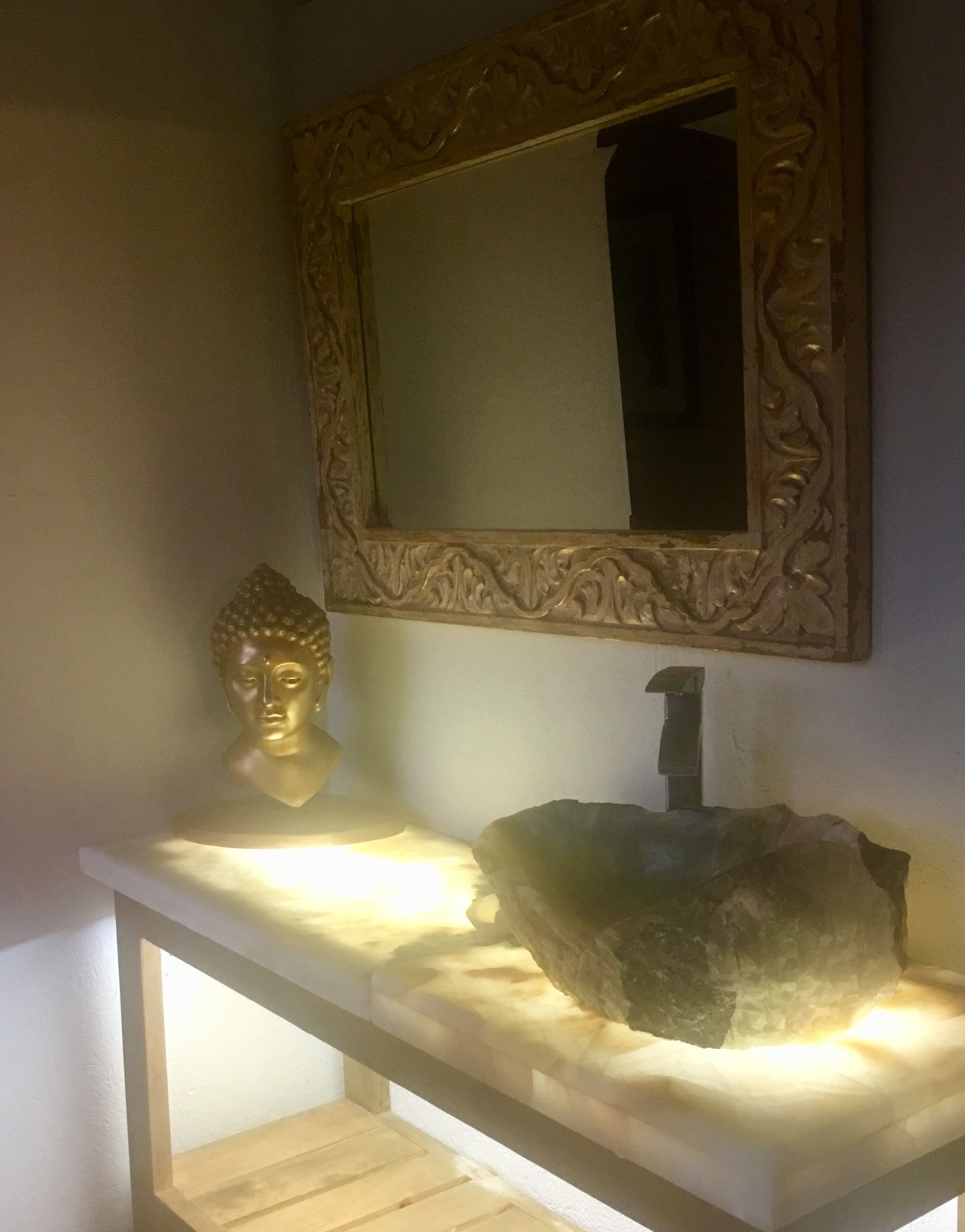 Een badkamer met een witte wastafel, een spiegel met gouden lijst, een Boeddha-beeld en een steen.