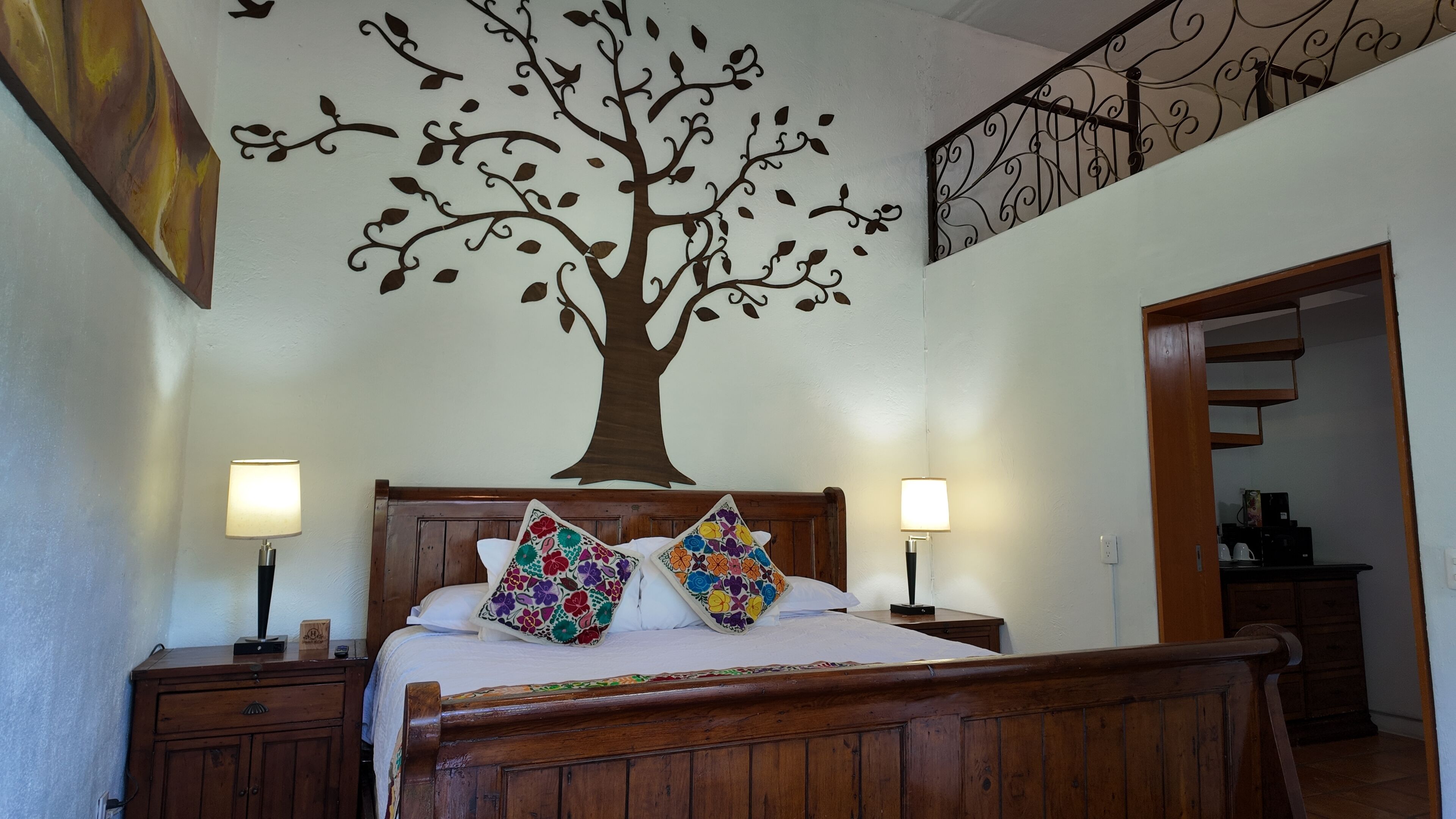 A bedroom with a wooden bed, two bedside lamps, a wall mural of a tree, and a staircase leading to another room.