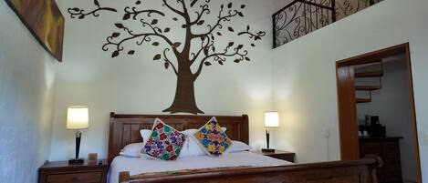 A bedroom with a wooden bed, two bedside lamps, a wall mural of a tree, and a staircase leading to another room.