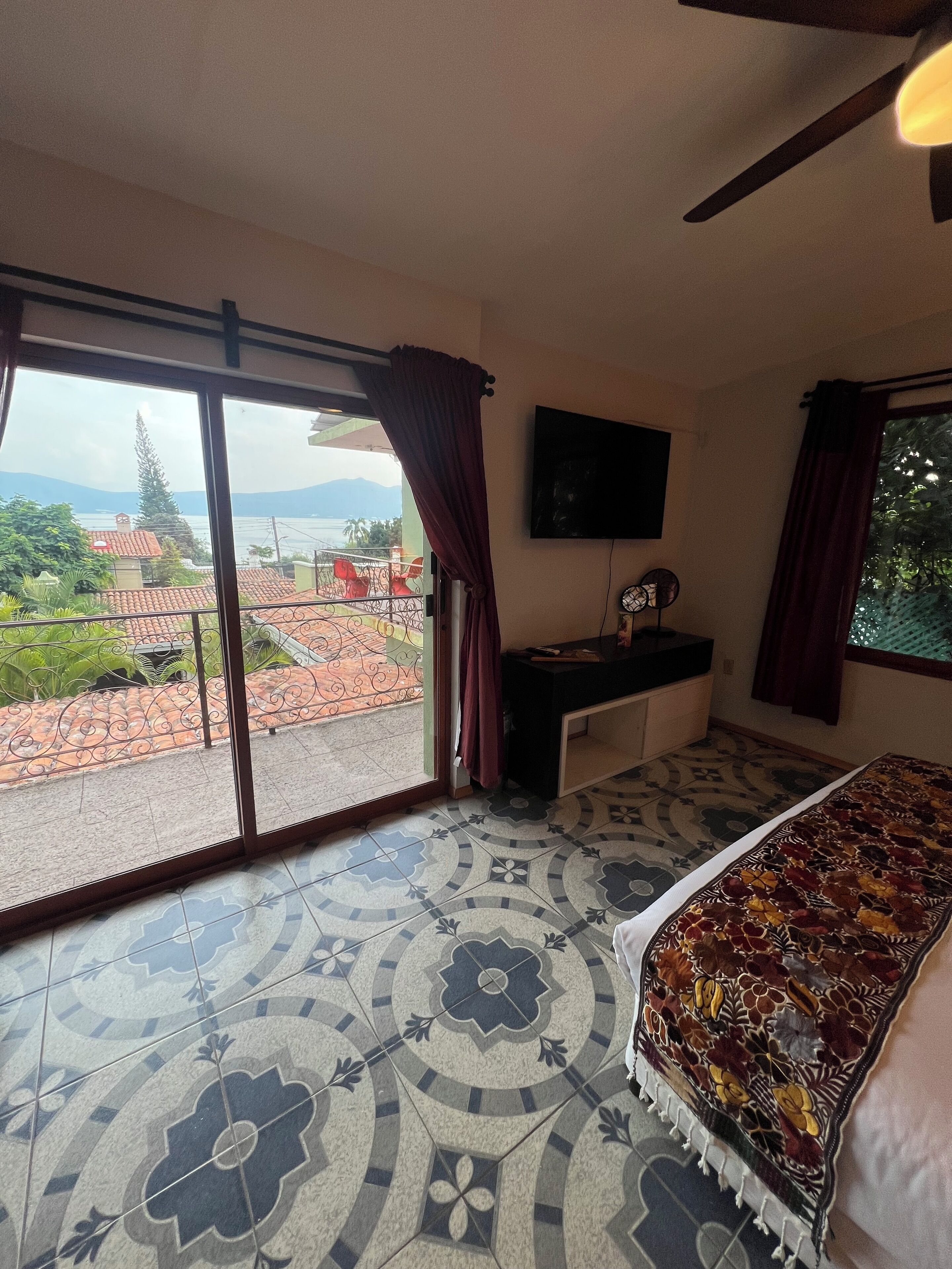 A bedroom with a large sliding glass door leading to a balcony, a bed with a patterned bedspread, a flat-screen TV mounted on the wall, and a small table with a fan.