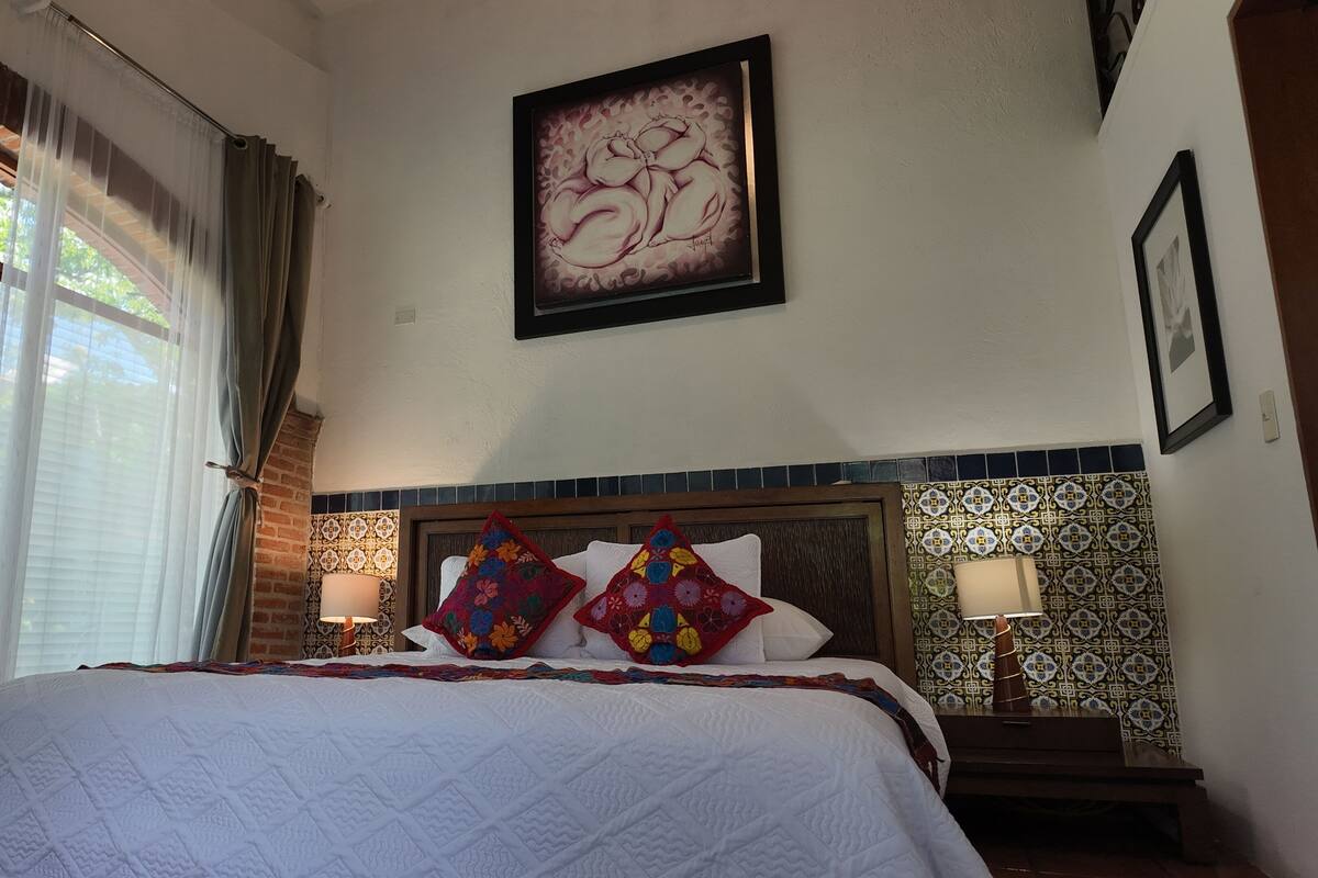 A hotel room with a large bed, a headboard with a patterned tile design, a painting on the wall, and a window with curtains.