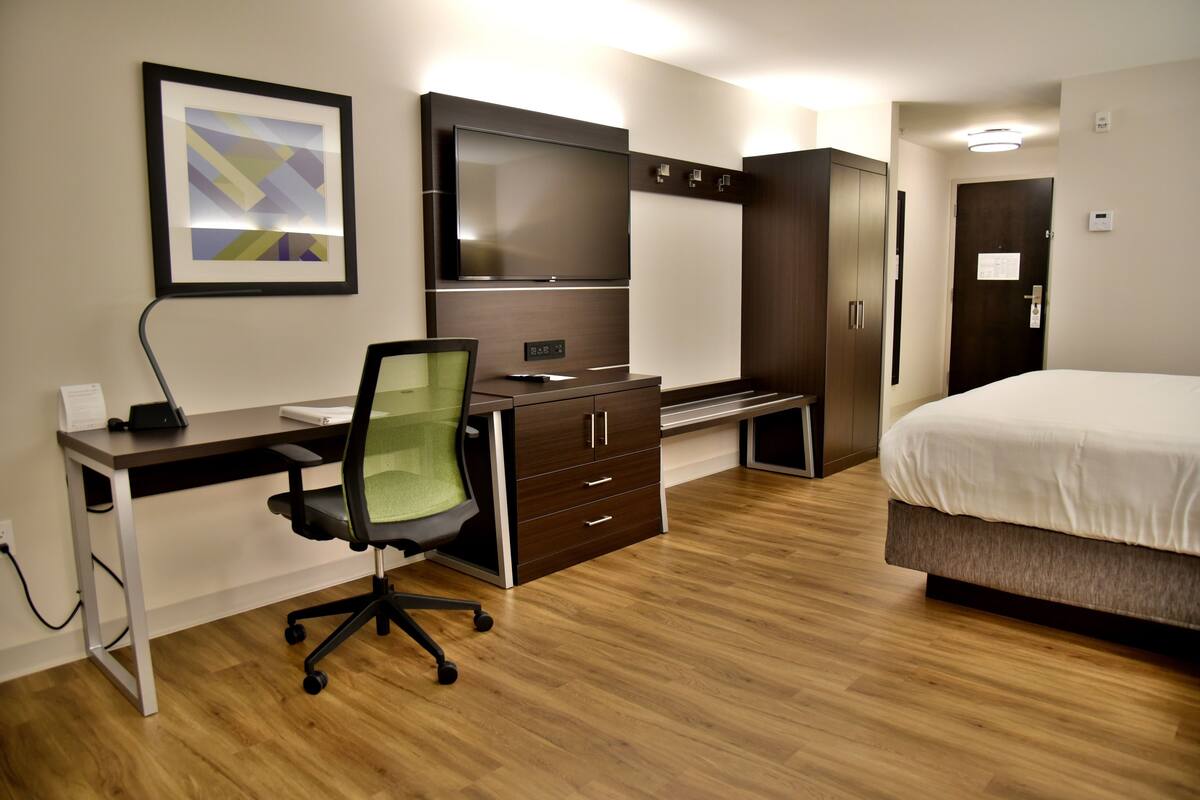 A modern hotel room with a wooden desk, a large flat-screen TV, a bed, and a wardrobe.