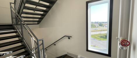 A stairwell with a metal railing, a window with a view of a parking lot, and a door with a red handle.