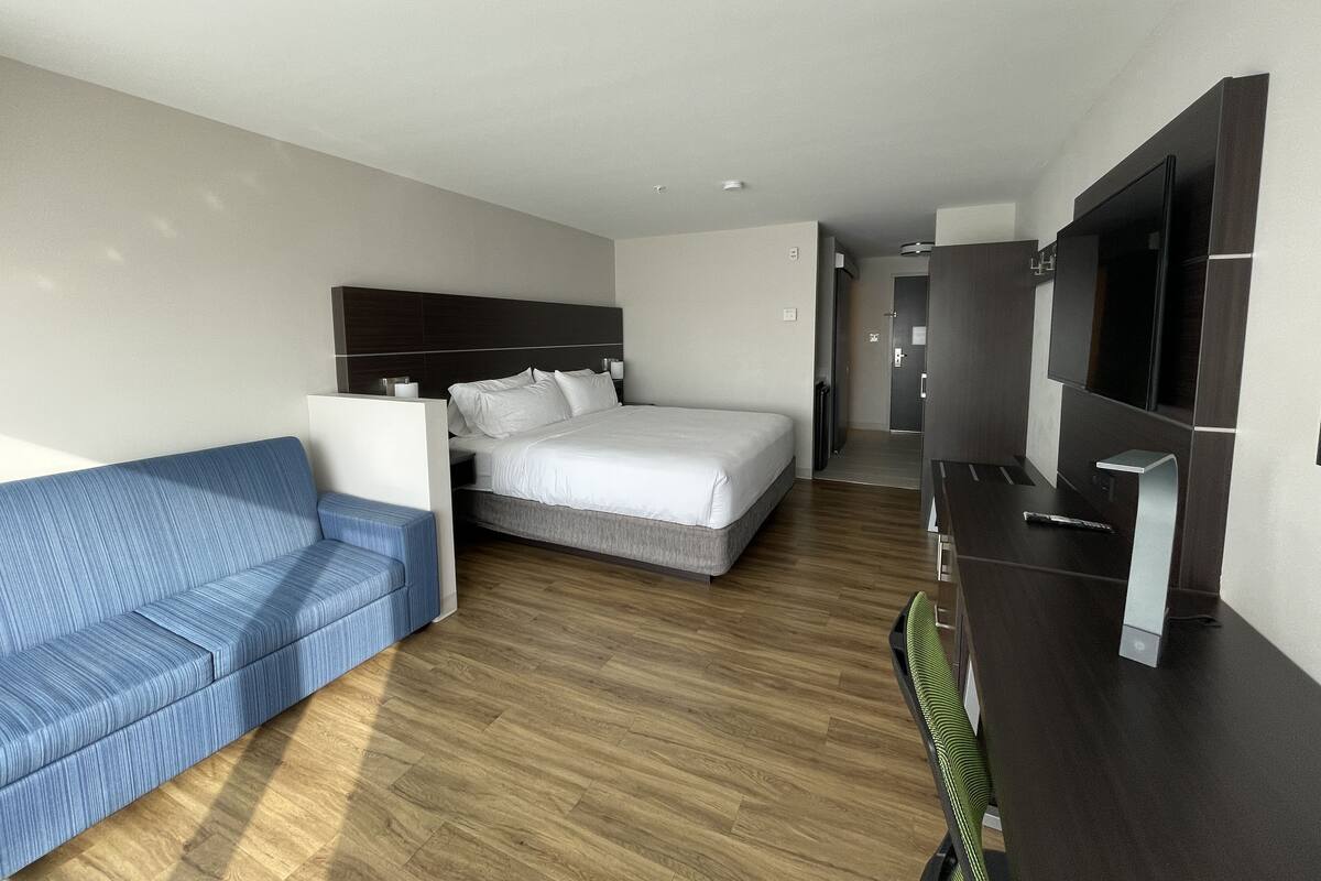 A modern hotel room with a bed, a blue sofa, a TV, and a wooden floor.