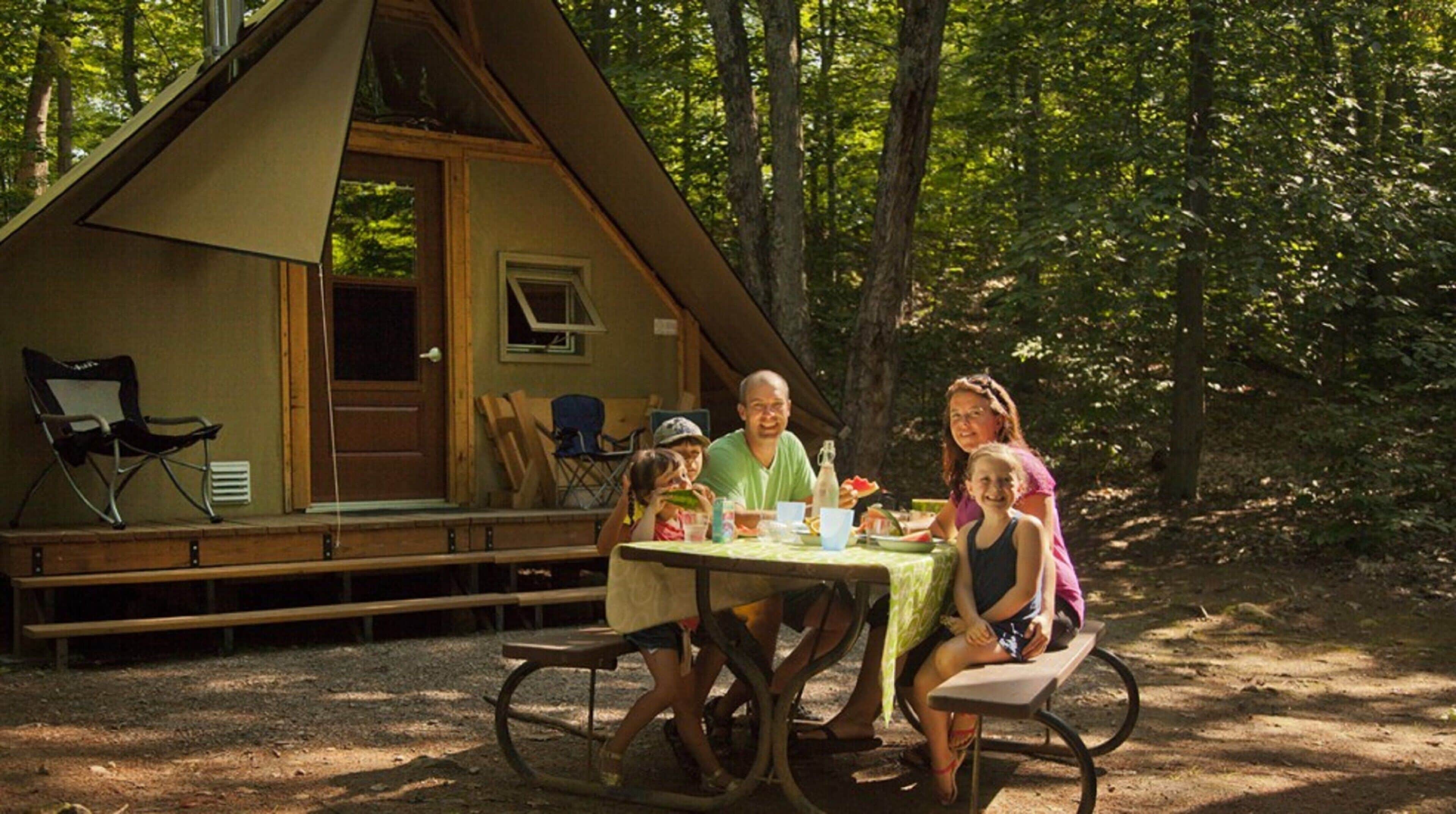 A family dining at a wooden table outside a cabin with a porch and a chair.